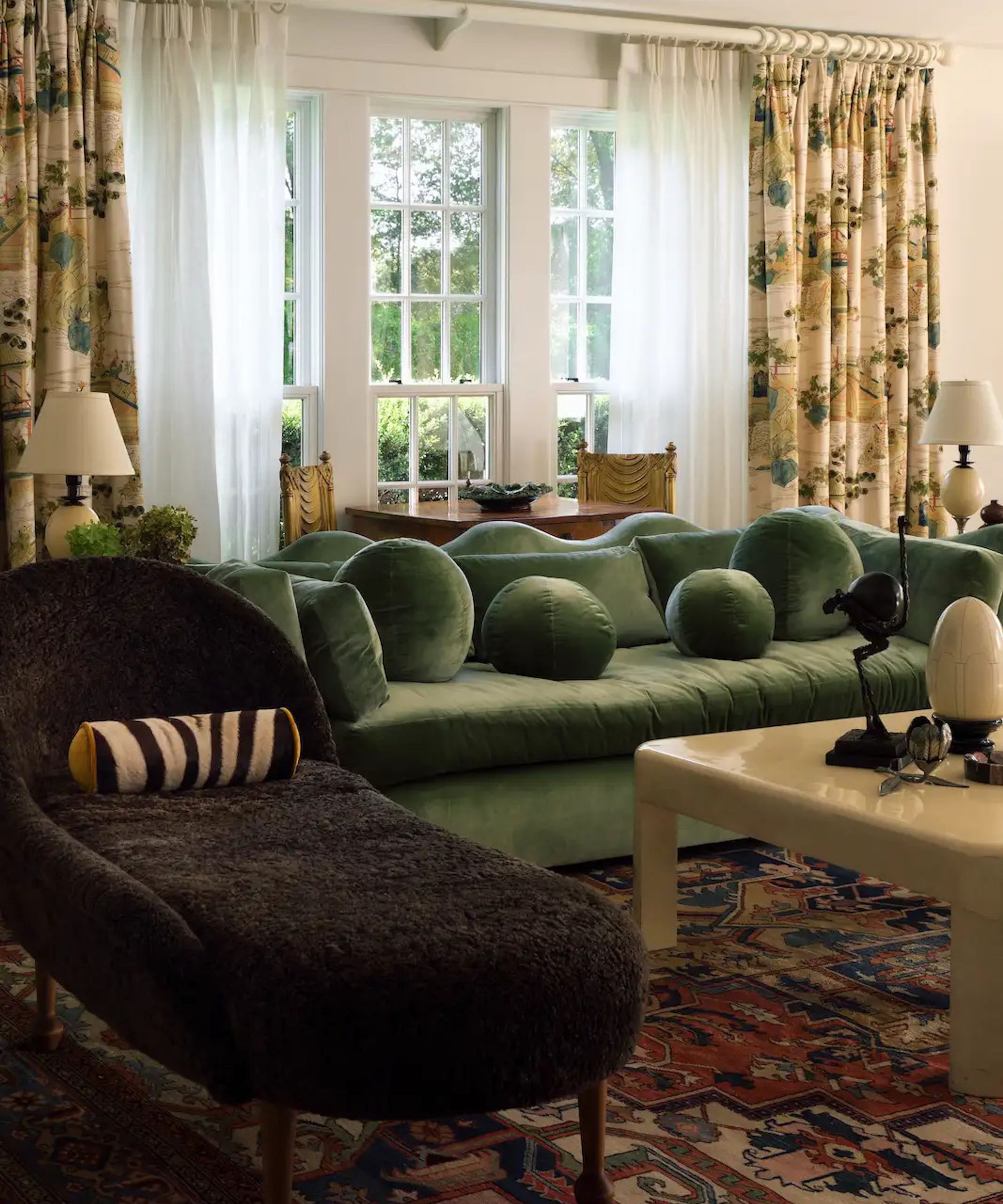 An eclectic living room featuring a long, moss-green velvet sofa with spherical pillows and a dark, textured chaise lounge. The room is decorated with a vibrant patterned rug, landscape-print curtains over sheer panels, and three large windows overlooking lush greenery