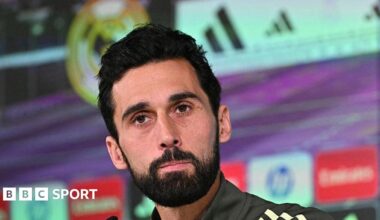 Alvaro Arbeloa speaks at a Real Madrid news conference, wearing a largely black club training top and with sponsorship logos in front of and behind him