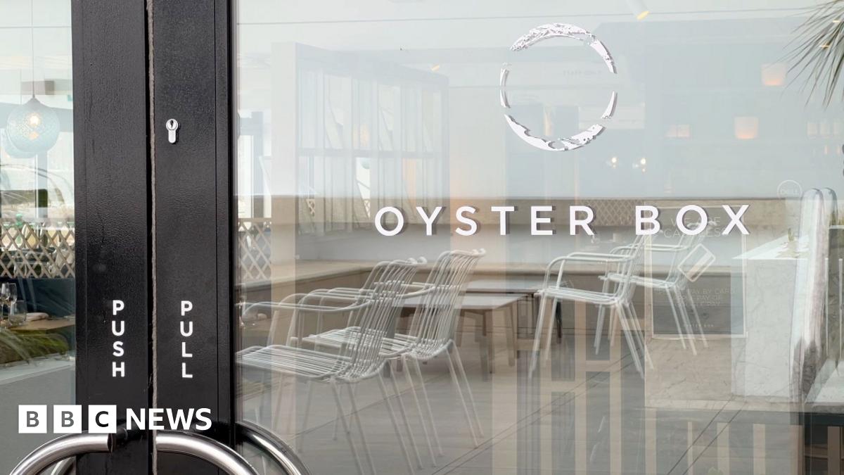 A glass door to a restaurant, with a black frame. On the glass, it says 'Oyster Box'. Above the words is a hollow white circular logo. Reflected in the glass, white metallic chairs are visible. On the black door frame are the words 'push' and 'pull' in white writing.