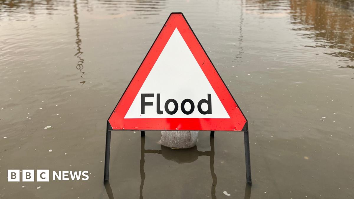 A red and white flood warning sign on a flooded road. The word flood is written in black letters.