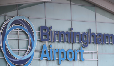 Birmingham Airport is written on the side of a building with a fogged glass exterior. The words are in dark blue and light blue lettering and there is a circular symbol to the left of the words.