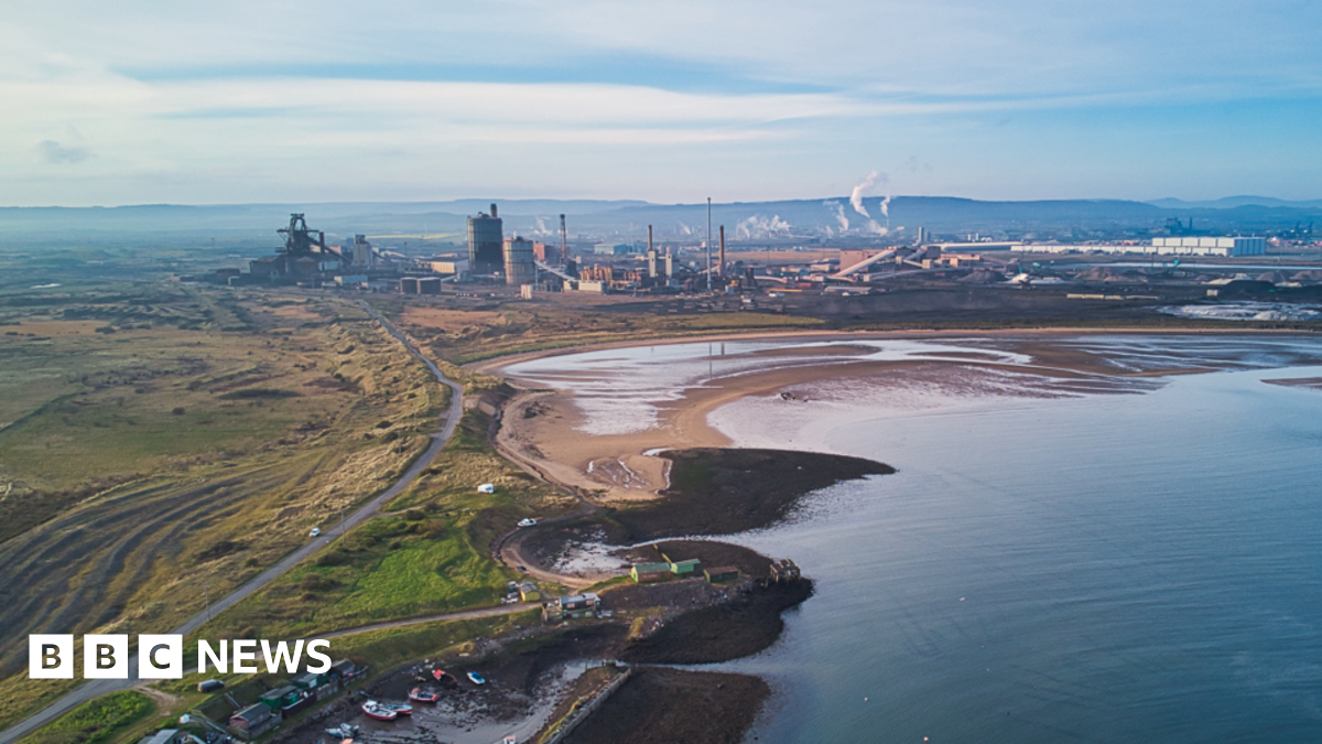 Overhead shot of coastal area, and a large industrial complex with high buildings and chimneys backing onto a large sandy bay, with further industrial buildings to the left. To the right is a scrubby grassed area. Low rising hills can be seen in the background.