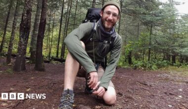 Alex is pictured in woodland wearing shorts and walking boots while kneeling on the ground