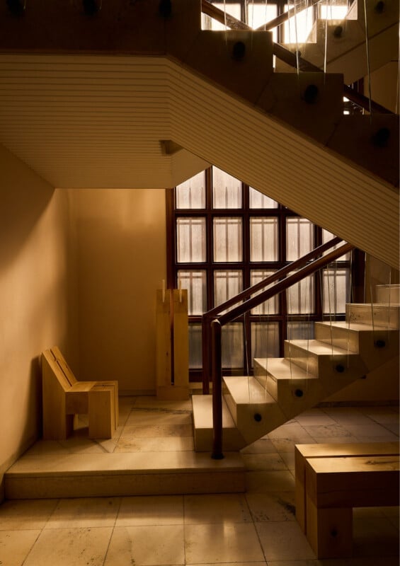 A sunlit stairwell with beige stone steps, glass railings, and modern wooden furniture, including a chair and a bench. Large grid windows let in soft natural light, casting shadows on the floor.