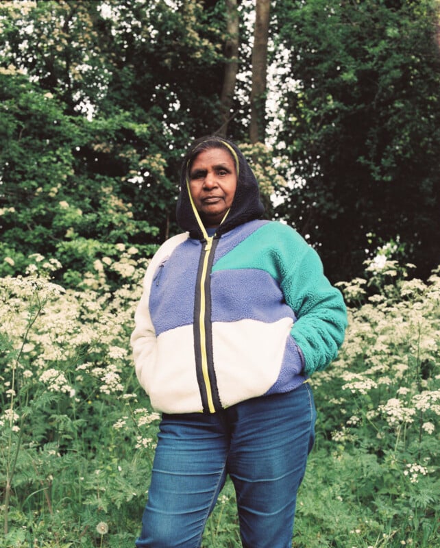 A woman stands outdoors in front of tall greenery and white wildflowers, wearing a hooded, color-block jacket and blue jeans, with her hands in her pockets, looking confidently at the camera.