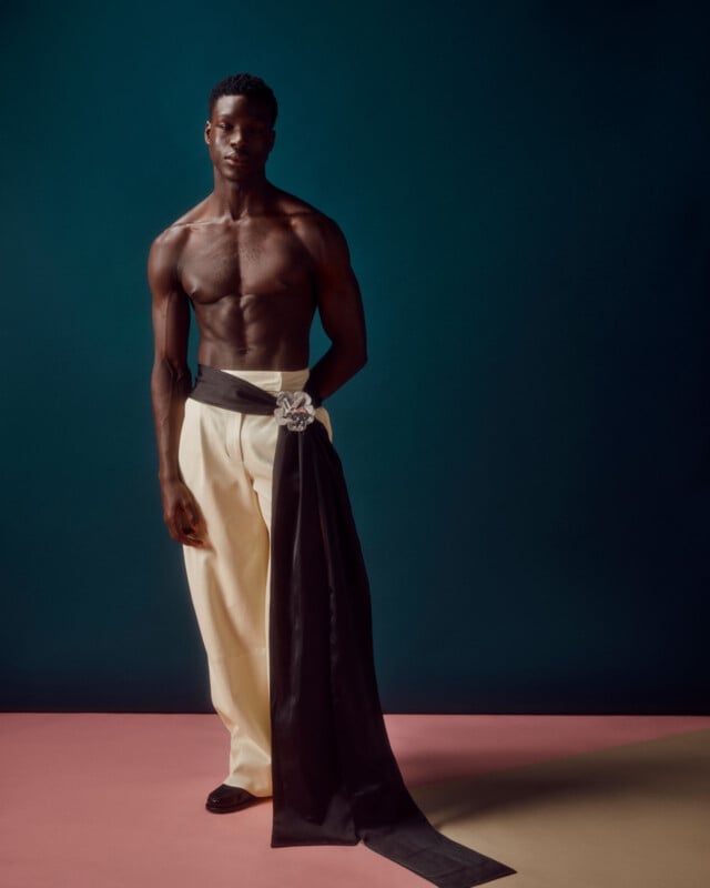 A shirtless man stands against a dark background, wearing high-waisted cream pants with a long, black sash featuring an ornate brooch at the waist. He poses with one arm relaxed and the other in his pocket.