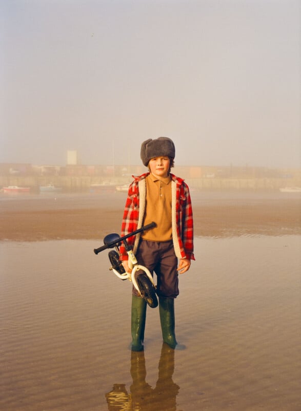 A child in a plaid jacket, fur hat, and green boots stands on wet sand, holding a small white bicycle. The background is misty, with water, boats, and a distant, hazy shoreline.