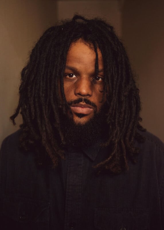 A person with long, dark dreadlocks and a beard looks directly at the camera, wearing a dark button-up shirt, standing against a softly lit neutral background.