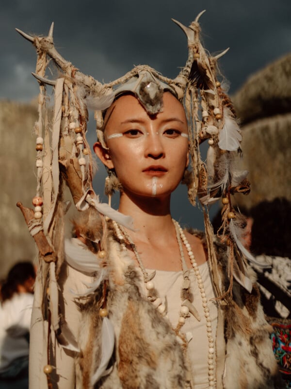 A person wears a headdress decorated with feathers, beads, bones, and fur, along with white face paint markings, standing outdoors with a dramatic sky and rocks in the background.