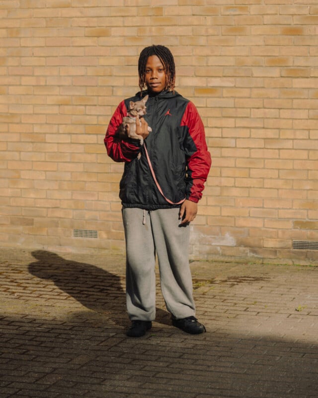 A young person in a black and red jacket and gray sweatpants stands on a paved area in front of a brick wall, holding a small gray dog on a pink leash.