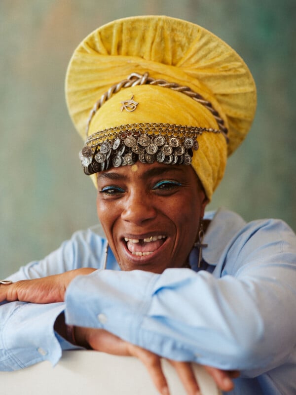 A joyful person wearing a bright yellow headwrap adorned with silver coins and jewelry, smiling widely and leaning forward with arms crossed, dressed in a light blue shirt. The background is softly blurred.