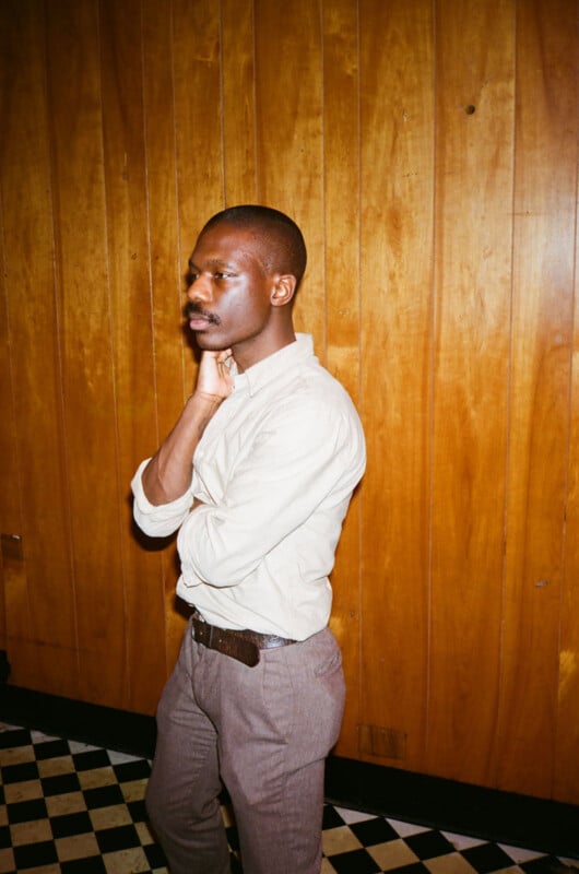A person wearing a light beige shirt and brown pants stands indoors against a wood-paneled wall with a black-and-white checkered floor, resting one hand on their neck and looking thoughtfully to the side.