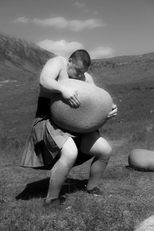A muscular person wearing a kilt and knee-high socks lifts a large stone outdoors on a grassy, mountainous landscape under a partly cloudy sky.