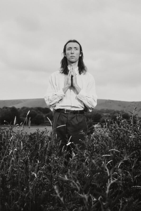 A person with long hair stands in tall grass with hands pressed together in a prayer gesture, looking upward. They wear a light-colored shirt and dark pants. Rolling hills and cloudy skies are in the background. The image is in black and white.
