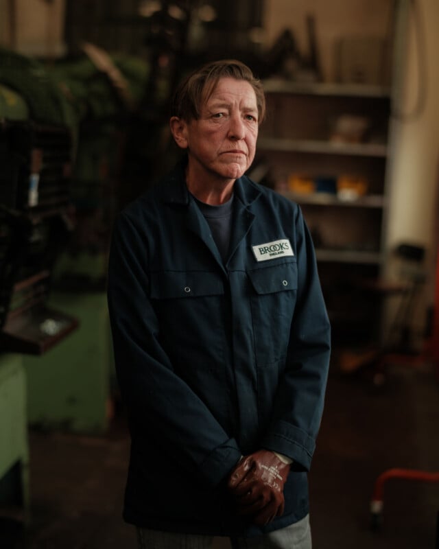 A person wearing a navy blue work jacket with a "Brooks" name tag stands indoors, hands clasped, in a workshop with shelves and machinery in the background.