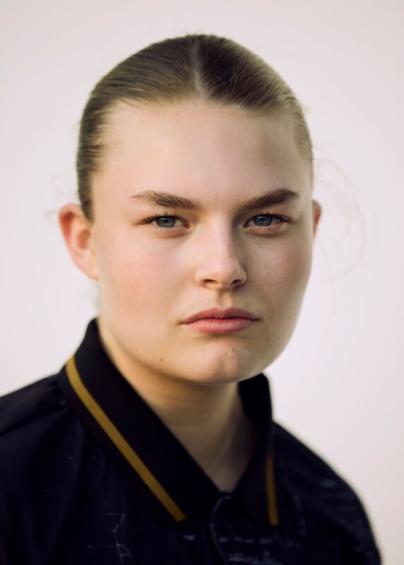 A person with light skin, blue eyes, and light brown hair pulled back is looking seriously at the camera. They are wearing a black collared jacket with a yellow stripe. The background is plain and light-colored.
