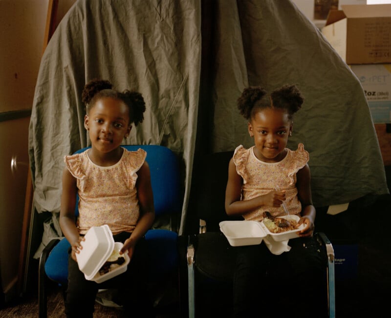 Two young girls with puffy hair sit on blue chairs, holding takeout containers with food. They wear matching pink ruffled tops and black pants. A gray sheet drapes behind them, with a cardboard box partially visible to the right.
