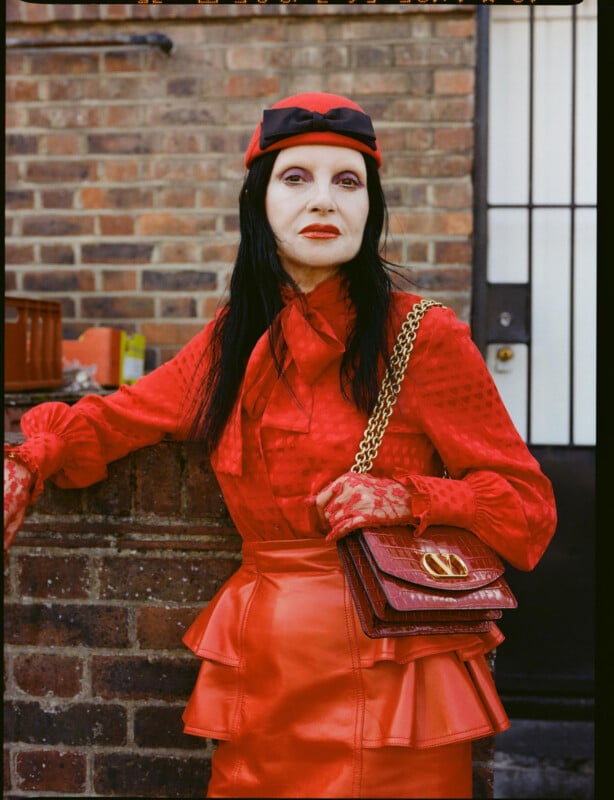 A woman with pale skin and long black hair leans against a brick wall. She wears a bright red outfit, including a sheer blouse, a leather skirt, a small hat, lace gloves, and a red handbag with a gold chain.