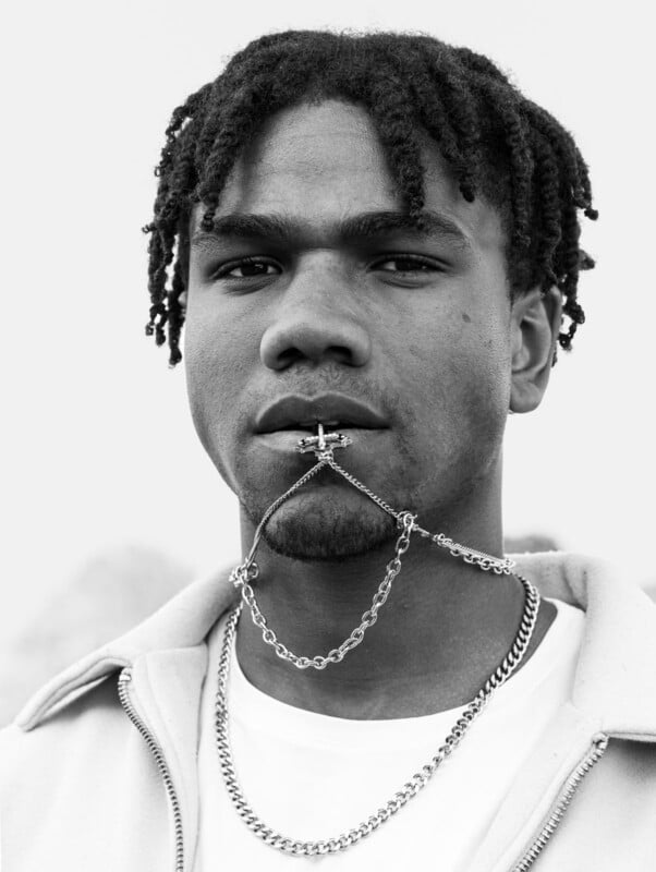 Black and white portrait of a young man with short twisted hair, holding a cross pendant in his mouth attached to a chain that wraps around his chin, wearing a light jacket over a white shirt and another chain necklace.
