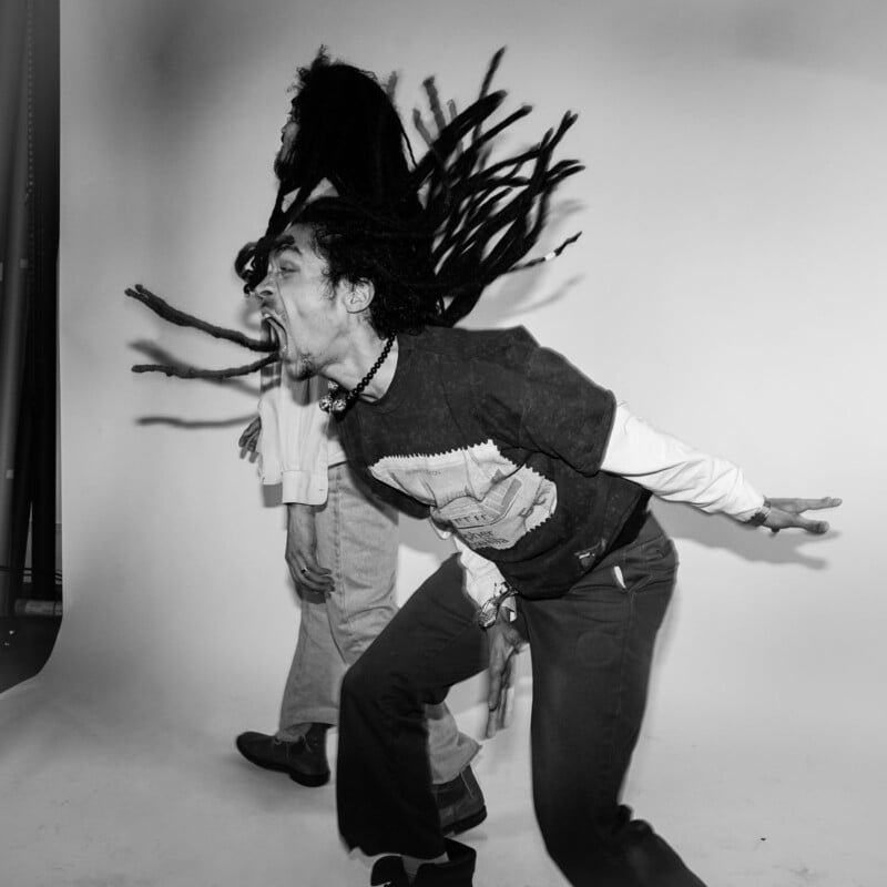 A person with long dreadlocks and an open mouth leans forward energetically, as if shouting or singing, while another person with dreadlocks stands in the background against a plain studio backdrop. Black and white photo.