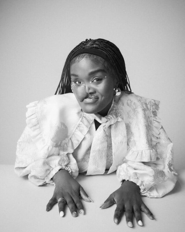 A woman with long braided hair, wearing a textured, ruffled blouse, rests her hands on a table and looks at the camera with a calm expression in a black and white portrait.
