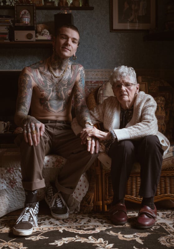 A heavily tattooed young man with piercings sits smiling next to an elderly woman wearing glasses and a white cardigan. They hold hands, seated on a patterned sofa in a cozy, vintage-style living room.