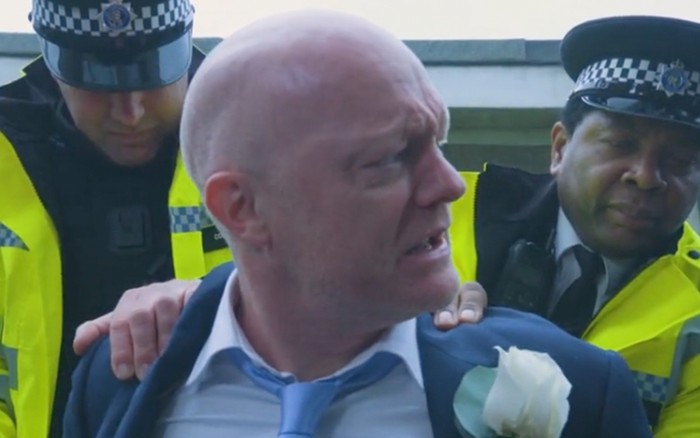 Jake Wood as Max Branning in a groom suit in close up and in side profile looking distressed as two police officers restrain him from behind in EastEnders.