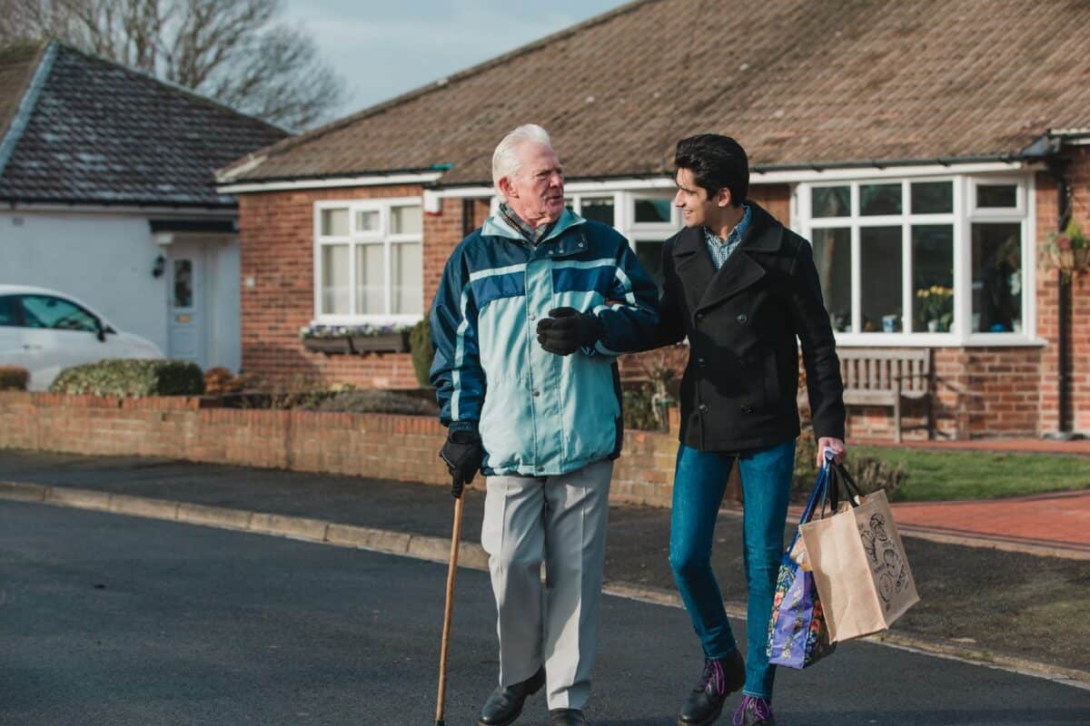 Teenage boy is walking back from the shop with his grandparent. He is carrying the shopping bag and they are linking arms.