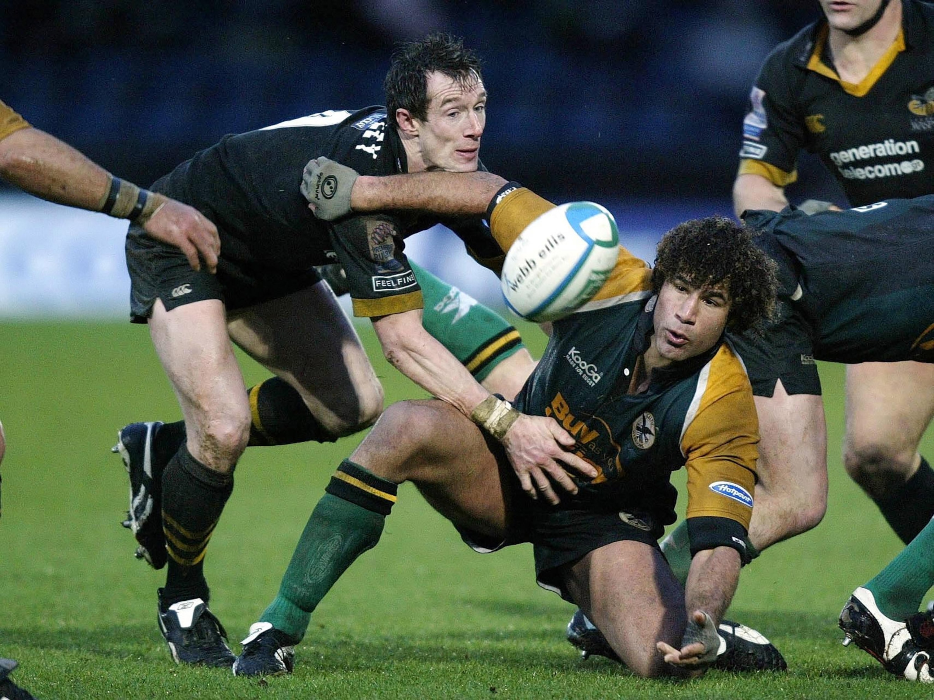 Two men on green turf in ruby uniforms reaching for an airborne rugby ball