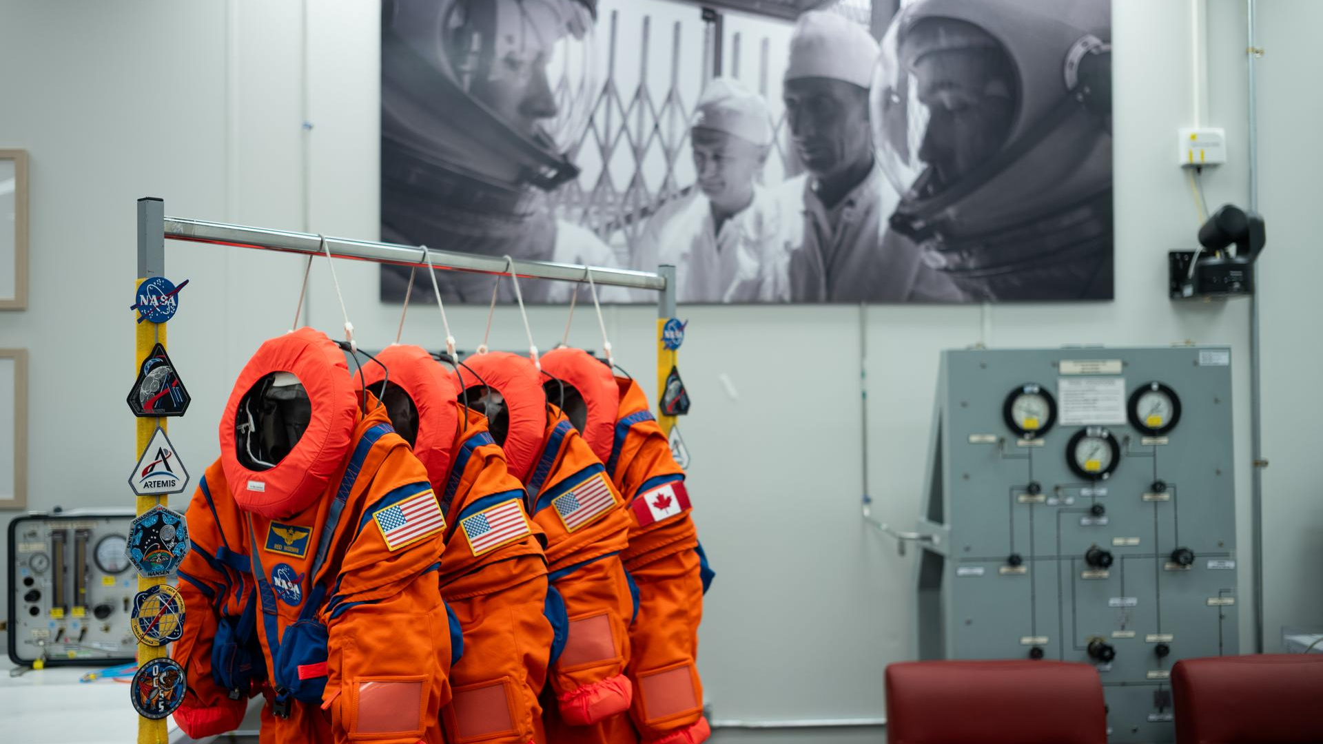 Four bright orange spacesuits with the circular NASA logo and American and Canadian flags on the arms hang on a rack in the middle of an empty room with chairs to the right and an image on the back white wall of two astronauts looking at each other