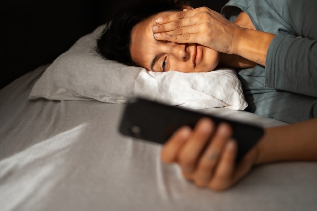 Woman protecting eyes from sunlight while holding phone in bed