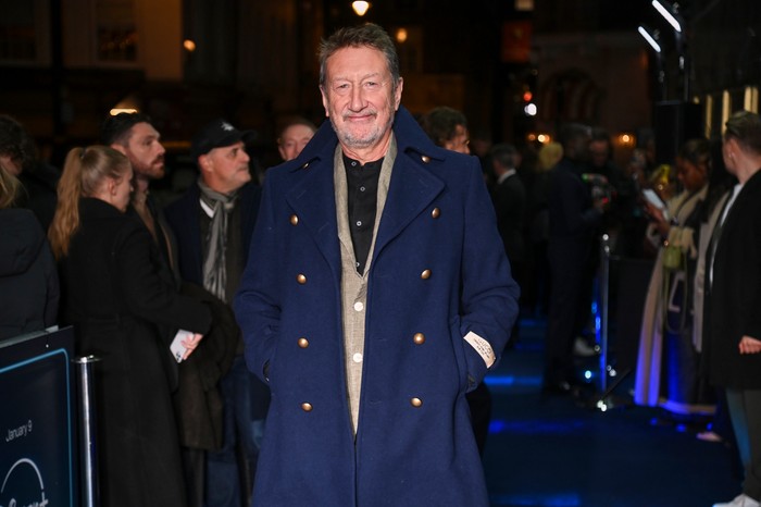 Steven Knight, wearing a blue coat and stood on a blue carpet, surrounded by people.