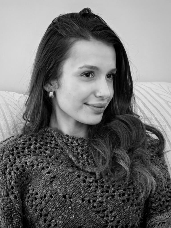 A young woman with long, wavy hair sits on a couch, wearing a textured sweater and small earrings, smiling softly and looking slightly to the side. The image is in black and white.