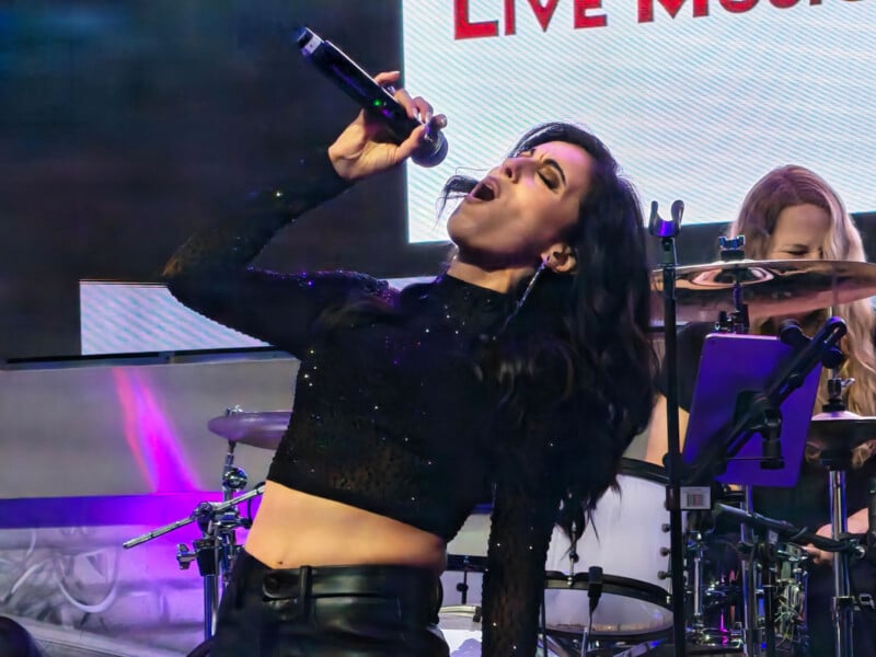 A woman in a black, long-sleeve sparkly top passionately sings into a microphone on stage, with a drummer and instruments in the background. Bright stage lights illuminate the lively performance.
