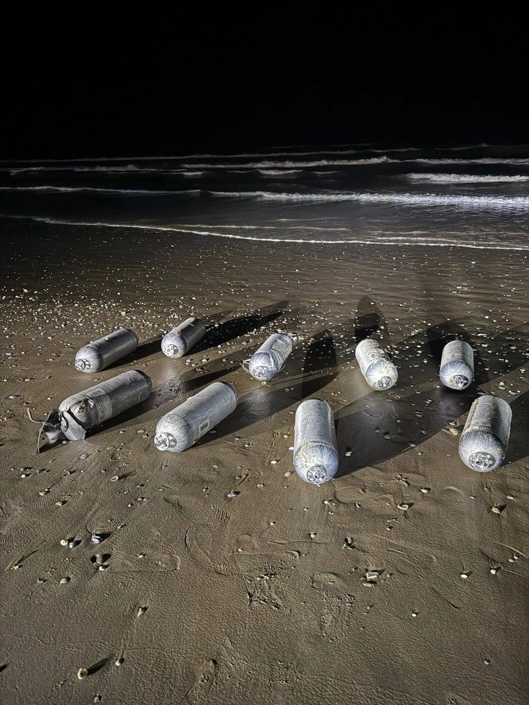 Nine large metal tanks wrapped in a plastic-like material lined up on a beach.