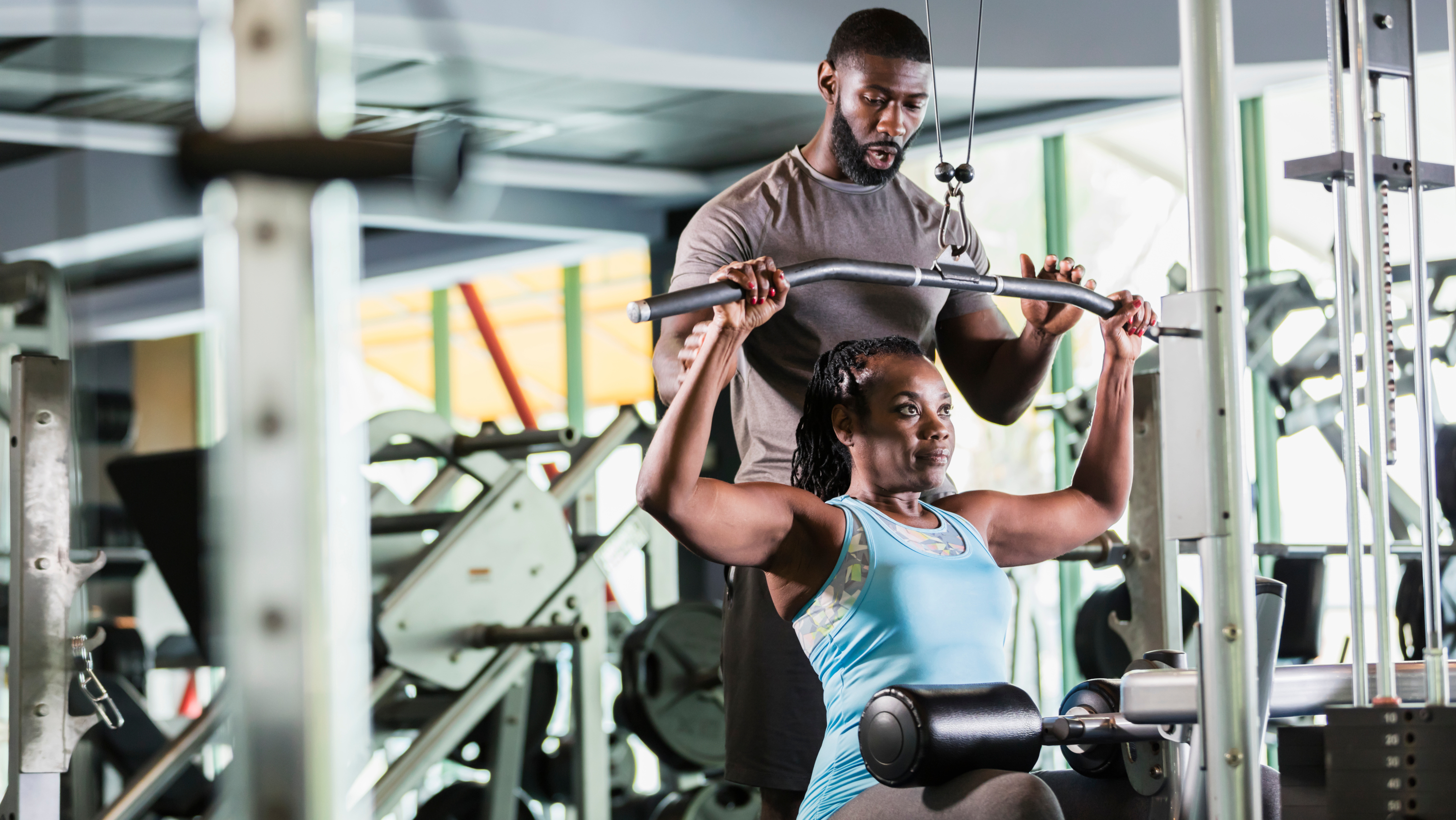 a photo of a woman working out on the lat pull down machine with a male trainer helping out