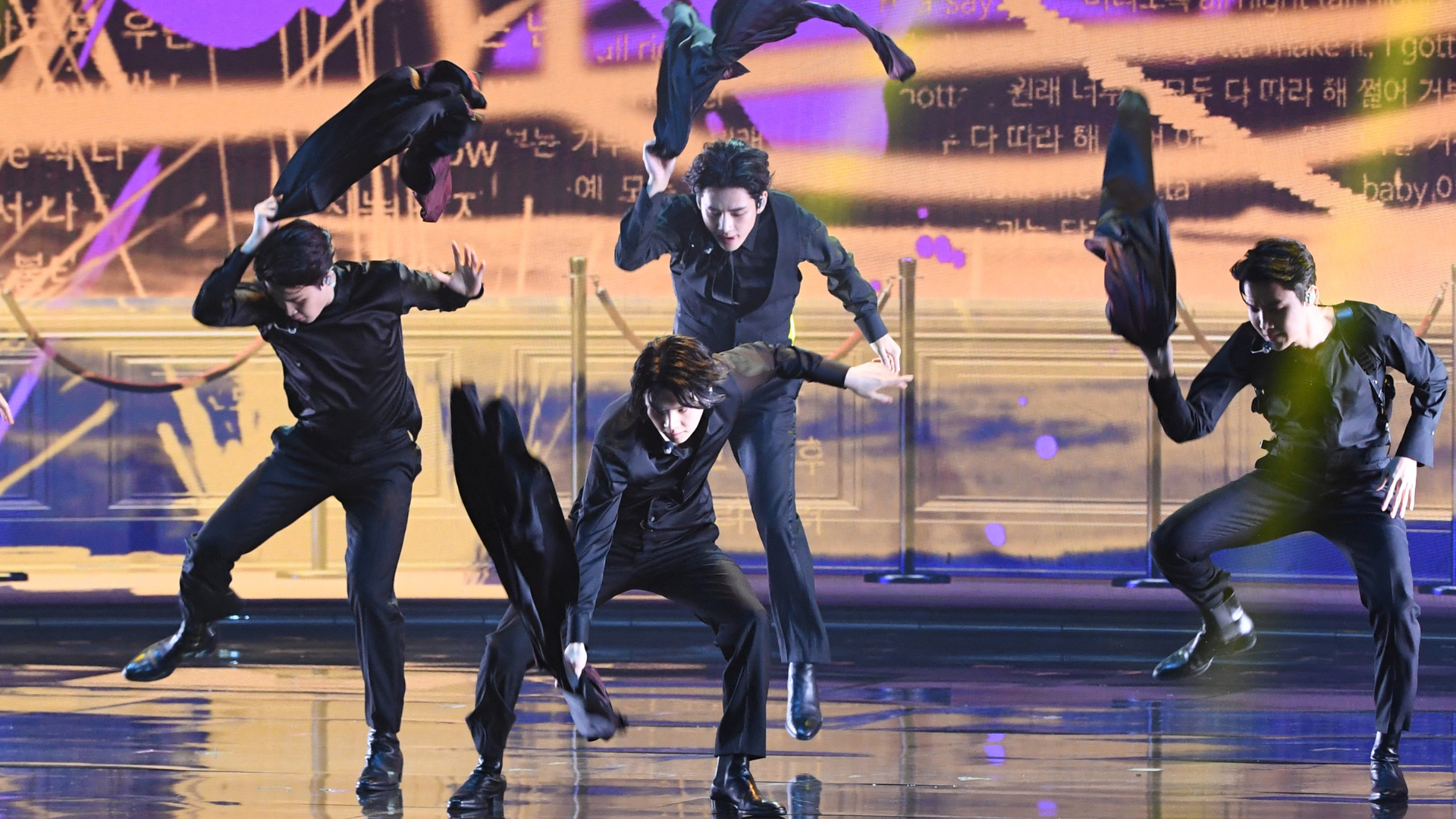 Four male dancers in black suits performing a choreographed routine on stage, holding or throwing black fabric.