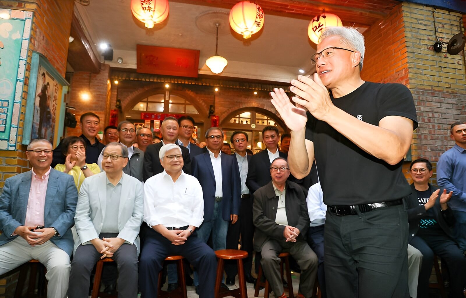 A group of people gathered indoors, with a man in a black t-shirt clapping at the front, surrounded by lanterns and a festive atmosphere.