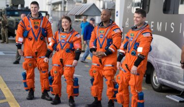 Four people in orange space suits stand on a road in front of a large van and look at a crowd off camera