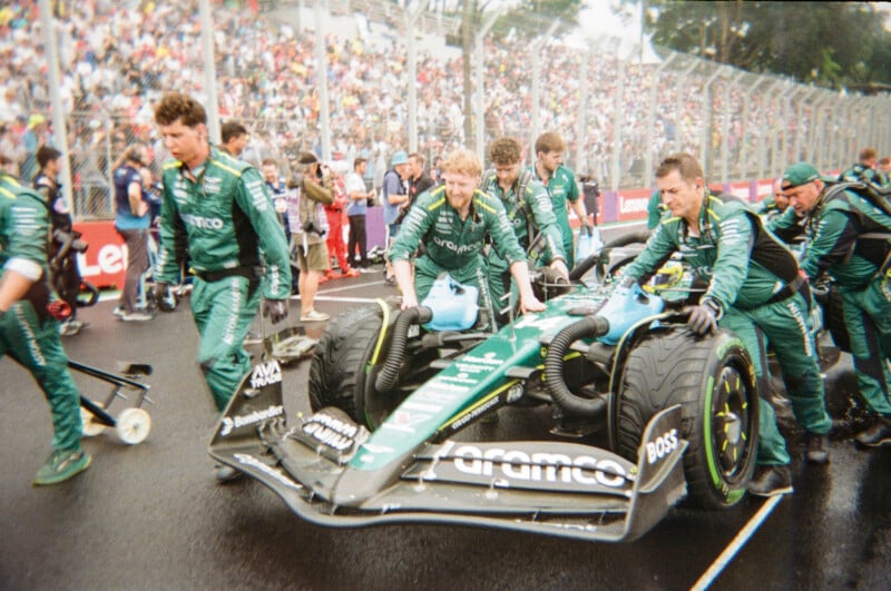 A Formula 1 team in green uniforms pushes their race car on a wet track with a large crowd in the stands behind a fence. The focus is on the crew and car during pre-race preparations.
