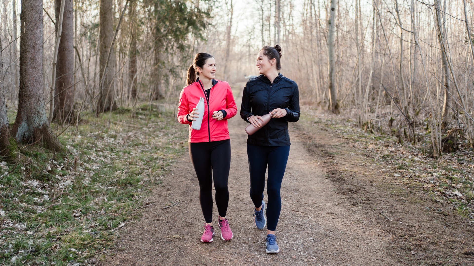 Two women walking and smiling together through the woods wearing activewear