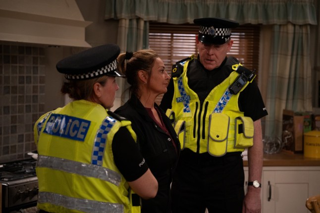 Moira standing between two police officers in the kitchen of Butlers farm in Emmerdale