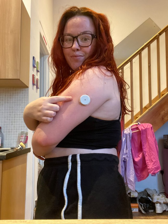 Bri Henderson, a type one diabetic, points at her continuous glucose monitor on her arm. She wears a blue strappy top and black and white trousers. Her hair is red.