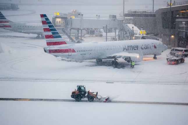 A snow removal machine is seen working while a Boeing 737 American Airlines passenger aircraft is parked at gate on the tarmac of LaGuardia airport in New York on January 25, 2026. A massive winter storm on January 24, 2026 dumped snow and freezing rain on New Mexico and Texas as it swept across the United States towards the northeast, threatening tens of millions of Americans with blackouts, transportation chaos and bone-chilling cold. Shoppers stripped supermarket shelves as the National Weather Service (NWS) forecast huge snowfall in some areas and possibly 