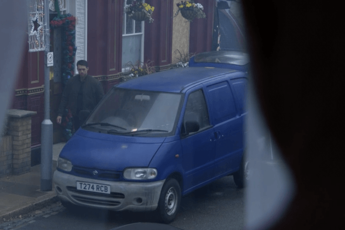 A man - portrayed by actor Stephen-Aaron Sipple - wearing a green jacket, walks past a blue van parked outside of The Queen Vic in the EastEnders flashforward episode. One of the pub's windows is boarded up.