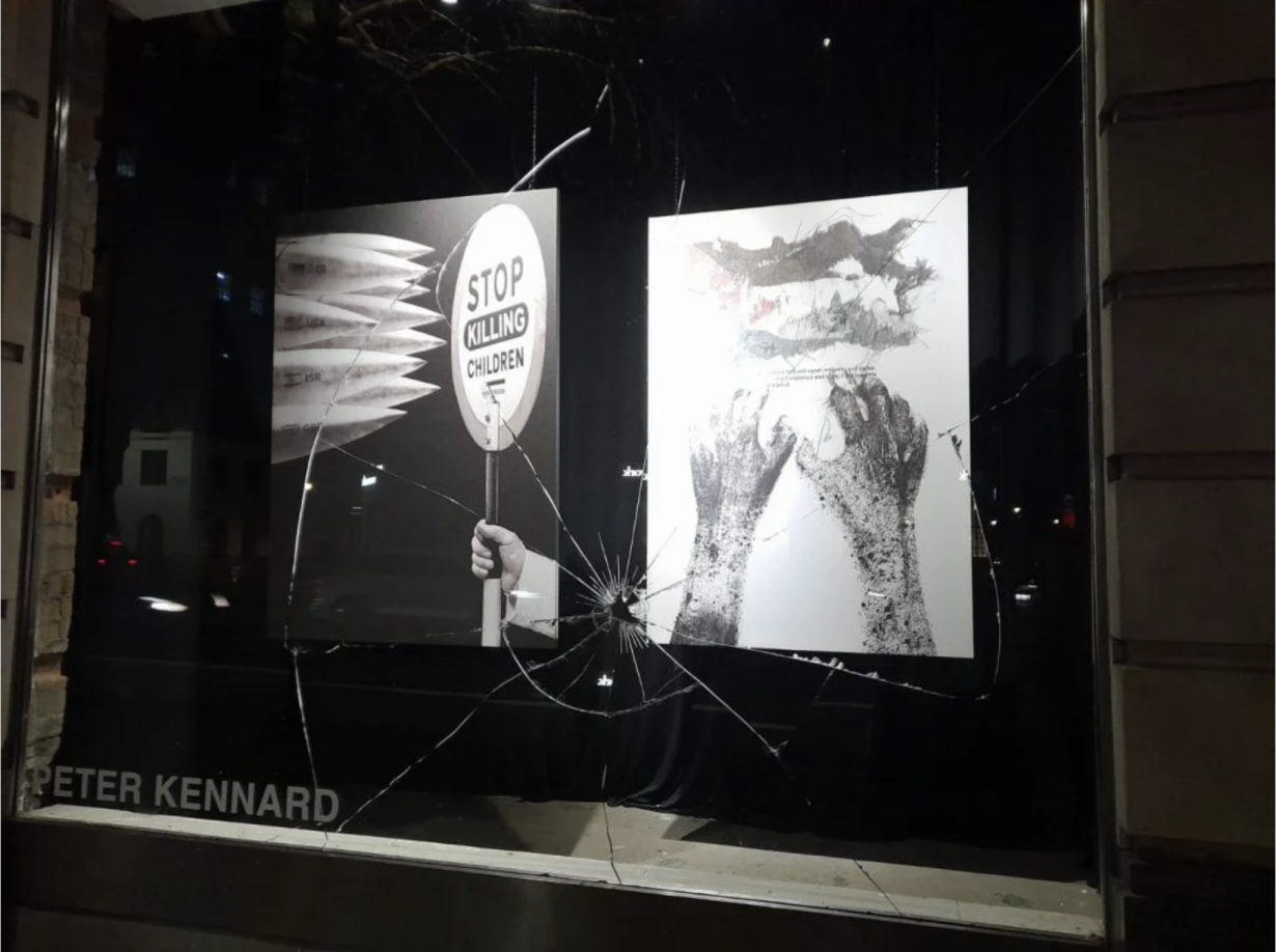 Smashed gallery window through which a work addressing Gaza by Peter Kennard is visible