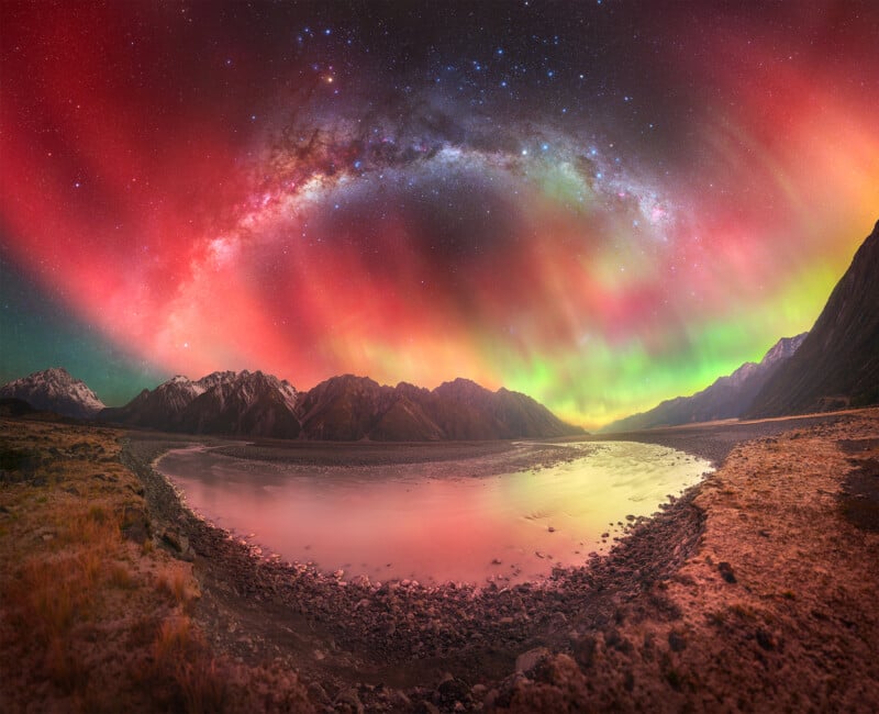 A vibrant night sky with colorful auroras in red, green, and purple hues arches above snowy mountains and a calm reflective lake, creating a stunning landscape scene.