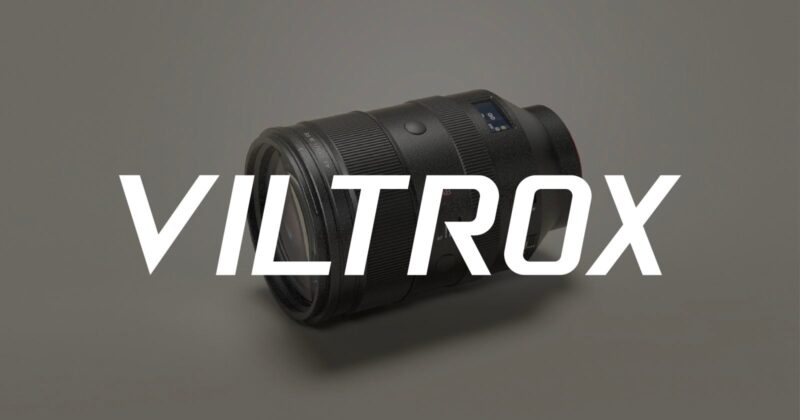 A black camera lens lies on a light gray surface with the word "VILTROX" in large, bold white letters displayed across the image.