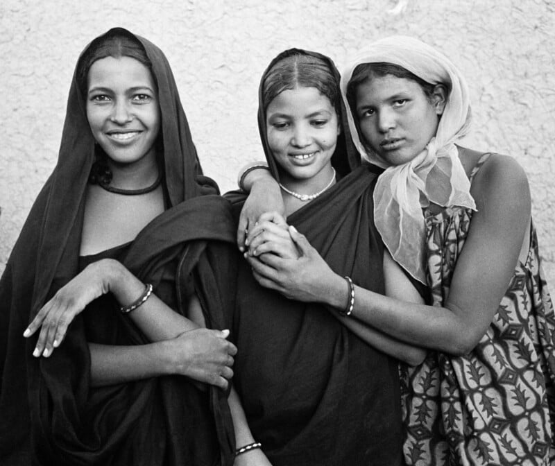 Three young women stand close together, smiling and embracing. They wear traditional clothing and headscarves, with bracelets on their arms, against a textured wall background. The photo is in black and white.