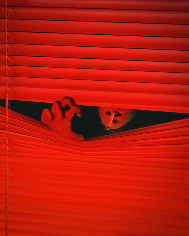A hand parts red blinds while a person with bright lipstick and a cigarette peers through the gap, their face partially hidden in shadow. Bold red lighting creates a dramatic, mysterious mood.